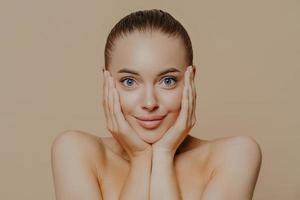 Headshot of young woman with surprised expression, touches face with both hands, has combed hair, natural makeup, healthy soft skin, stands against beige background. Facial treatment, cosmetology, spa photo