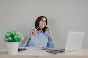 Attractive brunette female journalist arranges meeting via cell phone, works on new article on laptop computer, holds pen in hand as going to write some details in her notebook. Busy worker in office photo