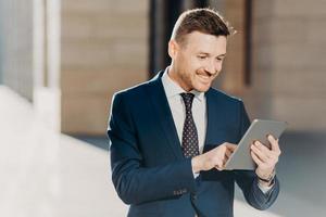 Succesful male entrepreneur in formal suit, holds modern tablet, checks documentation online, reads necessary information for making business report, uses free wifi connection. People and technology photo