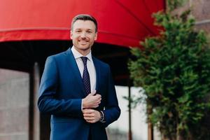 Positive male executive worker wears formal black suit, has positive expression, stands outdoor, waits for someone, being confident in achieving great success during his hard work and persistance photo