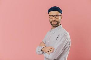 Studio shot of self assured bearded man with blue eyes, wears stylish hat and white shirt, keeps arms folded, models over pink background with free space on left side for your advertising content photo