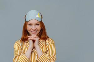 Pretty lady with ginger hair, keeps hands under chin, looks directly at camera, wears blindfold on head, striped yellow pyjama, has good rest, isolated over grey background, copy space for your text photo