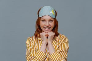 la foto de una mujer atractiva mantiene las manos debajo de la barbilla, tiene el pelo pelirrojo, una agradable sonrisa con dientes, usa ropa de dormir amarilla y máscara para dormir, mira directamente a la cámara, aislada sobre un fondo gris. despierta