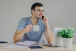 Handsome brunet male in round spectacles and casual t shirt, drinks hot beverage, has telephone conversation while works at office desk with laptop computer and documents. Communication concept photo