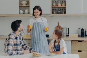 Lovely happy mother wears apron, holds two glasses of orange juice, talks to husband and daugher, prepares breakfast. Three membered family have dinner time at kitchen. Togetherness concept. photo