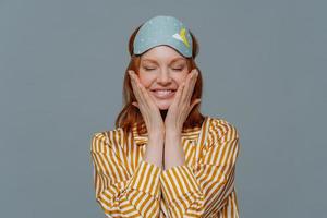 Pleased woman touches cheeks, has healthy freckled skin, smiles broadly prepares for sleep, wears striped pajama and sleep mask, closes eyes, isolated over grey background. Rest and lifestyle concept photo