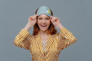 Ginger female model wears sleepmask and striped pajamas, awakes in good mood, realizes today is weekend, can stay in bed for long time, smiles positively, isolated on grey background. Sleeping photo