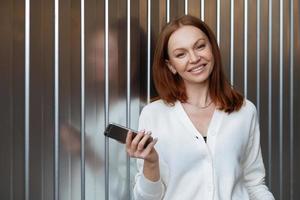 Satisfied female entrepeneur with toothy smile, wears white jumper, holds modern cell phone, sends text message, waits for business partner, stands indoor with free space for your promotion. photo