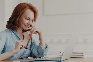 Pleased female model poses at coworking space, holds spectacles, smiles positively, uses wireless internet connection, reads information, has online communication, works on creating web project photo