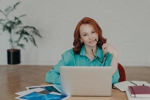 hermosa mujer pelirroja trabaja lejos de casa, se sienta frente a la computadora portátil termina de trabajar en el proyecto piensa en agregar contenido interesante en su propio sitio web prepara la publicación para la página web foto