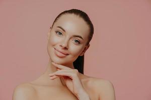 Charming beautiful woman with pony tail, touches perfect soft skin after cosmetology procedures, using beauty cream, stands with bare shouldes, poses against pink background. Natural as she is photo