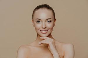 Cheerful young woman with clean glowing skin, keeps hand under chin, smiles gently, has well groomed body, combed hair, poses against beige background. Pure skin, beauty and wellbeing concept photo