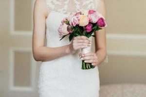 Wedding bouquet in bride's hands. Slim bride in white dress holding beautiful flowers in hands, going to marry, waiting for her future husband. Bridal bouquet. Wedding concept photo