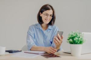 Pretty successful talented female copywriter blogs on smart phone, works on laptop computer, checks notification on modern electronic gadget, writes something in notebook with pen. Technology, job photo