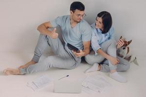 Photo of satisfied man wears spectacles and casual clothes, shows figures on calculator, demonstartes okay gesture, rejoices high profits, works together with female colleague, sit on floor.