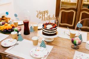 mesa festiva servida con pastel de cumpleaños, mesa llena de fruta, compota, platos vacíos y sombreros de fiesta. hermoso juego de mesa para fiesta de cumpleaños. composición festiva. concepto de celebración foto