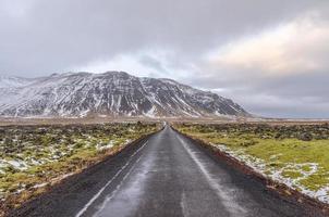 A moss-covered landscape of jumbled lava borders this narrow road in Iceland. The road continues on toward a snow dusted mountain ahead. photo