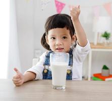 Little asian girl sitting at table in room, Preschooler girl drinking some milk with glass on sunny day, kindergarten or day photo