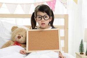 Child girl holding blank board on the wooden bed in her bedroom, Space for your text photo