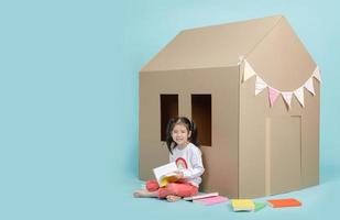 Asian little child girl reading a book with cardboard house isolated on blue long banner with copy space for your text, Homeschooling during the Coronavirus COVIC-19 concept photo