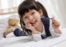 niña acostada boca abajo y sonriente, niña asiática feliz niña jugando en la cama de madera en su dormitorio, concepto de familia feliz foto