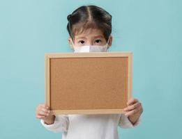 Asian little child girl wearing respirator mask to protect coronavirus outbreak and holding a blank board, New virus Covid-19 from Wuhan China concept, Empty space isolated on blue background photo