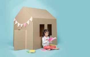 Asian little child girl reading a book with cardboard house isolated on blue long banner with copy space for your text, Homeschooling during the Coronavirus COVIC-19 concept photo