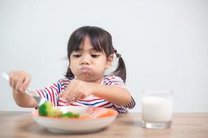 linda niña asiática comiendo vegetales saludables y leche para su comida foto