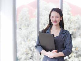 una joven empresaria asiática profesional viste un vestido gris y sonríe alegremente mientras trabaja y sostiene el portapapeles con confianza en el cargo. foto