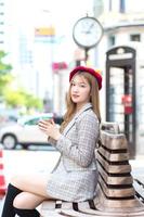 Asian beautiful woman who wears suit and red cap with bronze hair sits on chair in the city outdoors while holds coffee cup in her hand on a sunny morning. photo