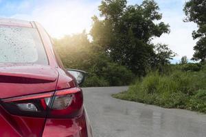 Rear side of red car with on the surface wet with raindrops. Driving on the asphalt road after rainy. Curved road in front and grass and green trees. Under the blue sky. Travel trips to the nature. photo