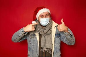 close up cute man in protective medical mask Santa hat dressed in winter clothes shows thumbs up with index finger on his face isolated on red studio background. concept - viruses, health preservation photo