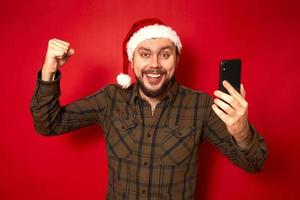 Portrait of crazy excited christmas father raising fist screaming eyes got news life holding cell phone wearing santa hat green shirt isolated over red background concept victory, holiday, technology photo