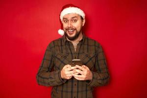 Wow, I got message from stranger. Fashionable guy in santa hat and green shirt, holds cell phone, gasps from surprisement, reads stunning news, poses over red wall with empty space for information photo