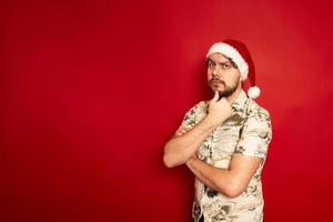 Puzzled confused young traveler tourist man in summer casual clothes christmas santa hat put hand on chin isolated on red studio background. Passenger traveling on holiday. Air flight journey concept photo