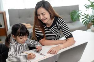 Niña pequeña asiática aprendiendo clases en línea en casa con su madre. el niño en edad preescolar usa una computadora portátil para hacer la tarea, la escuela en casa del maestro de la escuela por internet remoto digital con el apoyo de mamá. foto