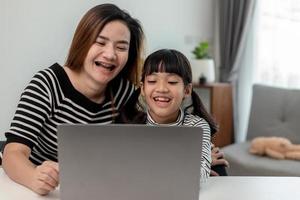 Asian little young girl kid learning online class at home with mother. Preschool child use laptop computer do homework, homeschool from school teacher by digital remote internet with support from mom. photo