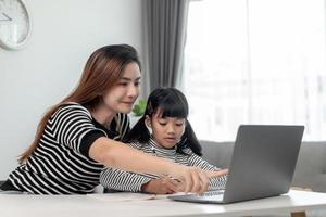 Asian little young girl kid learning online class at home with mother. Preschool child use laptop computer do homework, homeschool from school teacher by digital remote internet with support from mom. photo