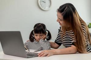 Asian little young girl kid learning online class at home with mother. Preschool child use laptop computer do homework, homeschool from school teacher by digital remote internet with support from mom. photo