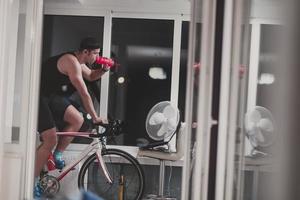 hombre en bicicleta en el entrenador de la máquina que está haciendo ejercicio en el hogar por la noche jugando al juego de carreras de bicicletas en línea foto