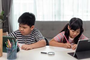 los niños y niñas asiáticos disfrutan del aprendizaje en línea tomando notas y usando tabletas en casa. foto