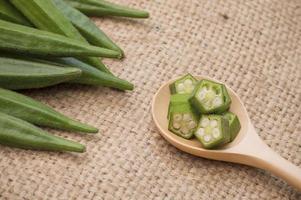 okra fresca sobre mesa de madera y rodajas de okra en cuchara de madera sobre fondo de tela de saco foto