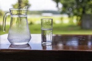 A jug of water and a glass of cold water are placed on a wooden table against a beautiful backdrop. photo