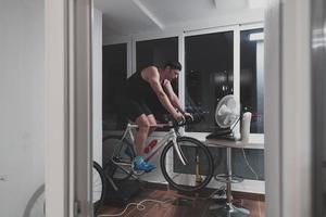 hombre en bicicleta en el entrenador de la máquina que está haciendo ejercicio en el hogar por la noche jugando al juego de carreras de bicicletas en línea foto