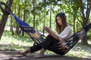 An Asian woman sits in a hammock while working with a tablet under a shady forest. photo