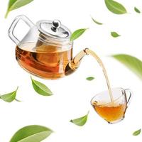 jarra que vierte té caliente en una taza de vidrio con hojas de té verde en el aire, productos saludables por concepto de ingredientes naturales orgánicos, espacio vacío en una foto de estudio aislada en fondo blanco