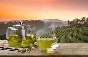 Cup of hot green tea and glass jugs or jars and reen tea leaf on the wooden table and the tea plantations background photo