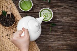 vierta a mano té verde humeante caliente en una taza de tetera y té de hierbas secas en la mesa de madera espacio vacío creativo plano, producto orgánico de la naturaleza para una salud saludable con estilo tradicional foto