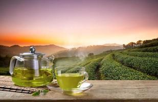taza de té verde caliente y jarras o frascos de vidrio y hojas de té reen sobre la mesa de madera y el fondo de las plantaciones de té foto