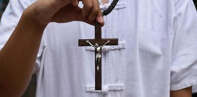 la cruz de madera y el rosario de madera se sostienen en manos de jóvenes católicos asiáticos rezando mientras rezan en el área del parque del templo. foto